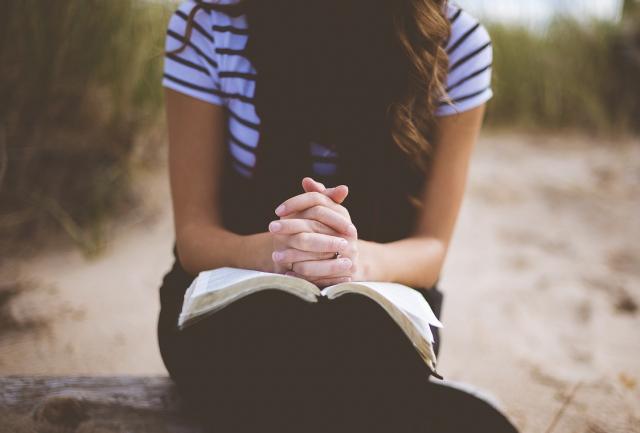 Tyttö istuu rannalla rukoilemassa avoin Raamattu sylissään. Kädet lepäävät Raamatun päällä.