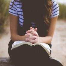 Tyttö istuu rannalla rukoilemassa avoin Raamattu sylissään. Kädet lepäävät Raamatun päällä.